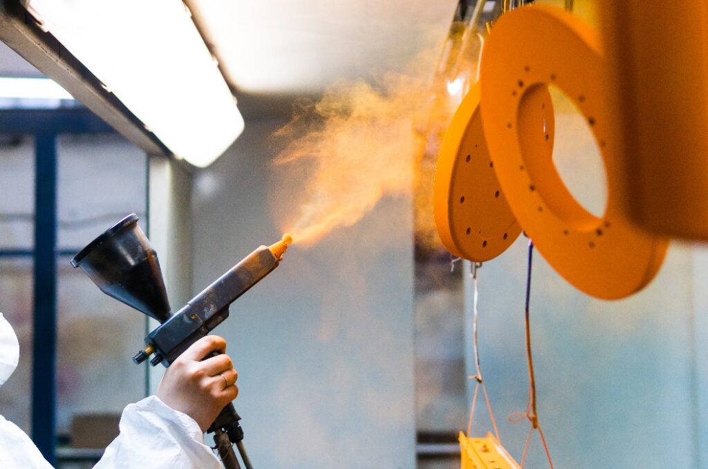 Pulverbeschichtung von Metallteilen. Eine Mitarbeiterin in Schutzkleidung sprüht mit einer Pistole Pulverlack auf Metallprodukte.